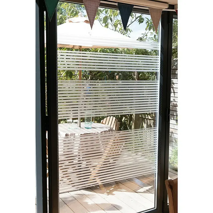 D-c-fix StaticStripes Glasfolie Ava - 200 x 45 cm, Transluzent, Statisch haftend - Nahaufnahme eines dekorierten Fensters mit horizontalen Streifenfolien, die ein verschwommenes Gartenbild mit Tisch und Sonnenschirm zeigen.