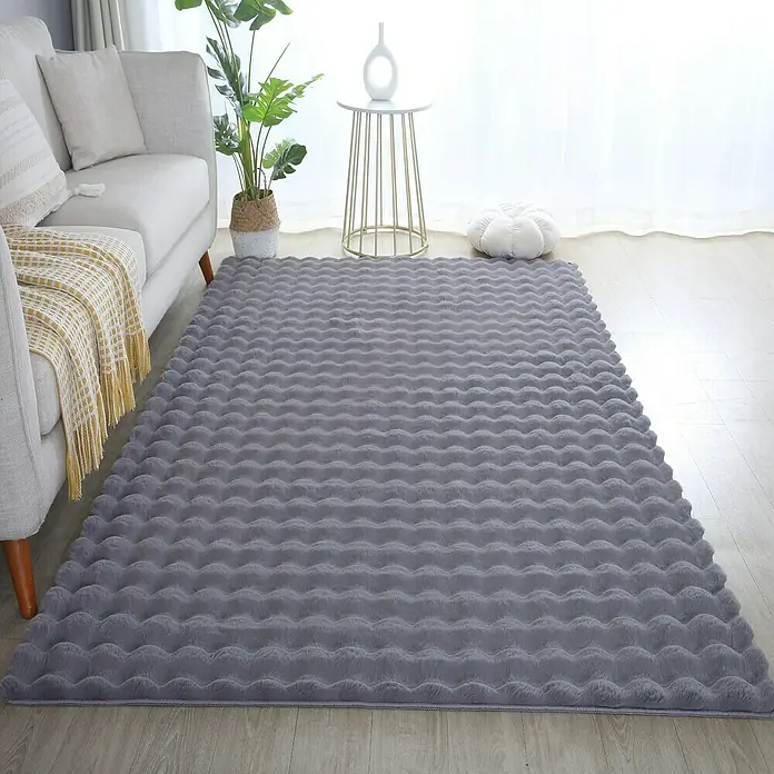 Fellteppich Ambiance - Grau, 150 x 80 cm, 100 % Polyester - Nahaufnahme eines weichen, grauen Teppichs mit gewellter Textur, der vor einem Sofa und einem Beistelltisch auf einem Holzboden liegt.
