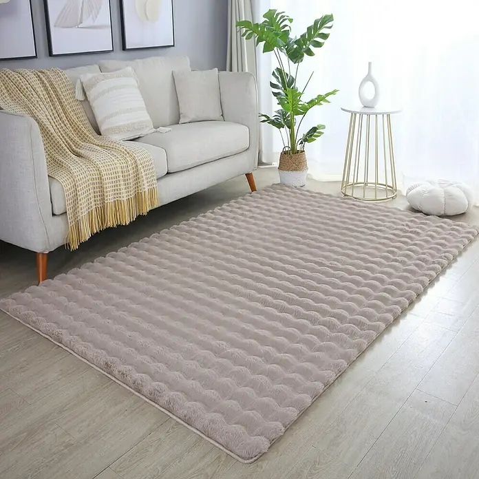 Fellteppich Ambiance - Beige, 200 x 140 cm, 100 % Polyester - Ein beiger, rechteckiger Teppich mit geriffelter Textur liegt in einem Wohnzimmer mit Couch, Beistelltisch und Zimmerpflanze.