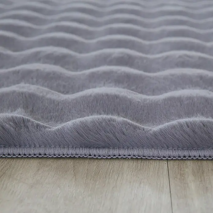 Fellteppich Ambiance - Grau, 150 x 80 cm, 100 % Polyester - Nahaufnahme eines grauen, gewellten Teppichs mit weicher Textur, der auf einem hellen Holzboden liegt. Der Teppich hat eine dekorative Bordüre.