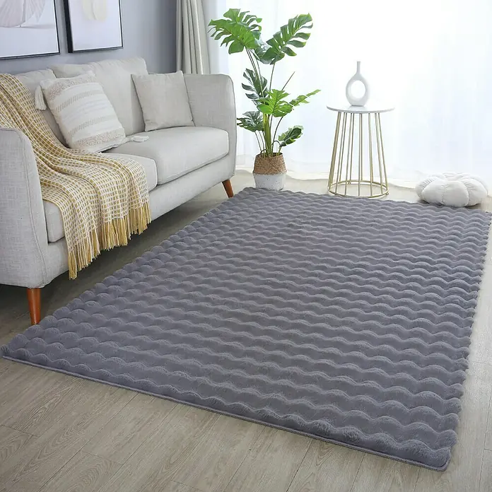 Fellteppich Ambiance - Grau, 150 x 80 cm, 100 % Polyester - Ein grauer, gewellter Teppich liegt im Wohnzimmer und sorgt für eine gemütliche Atmosphäre. Couch, Pflanze und Deko ergänzen den Raum.