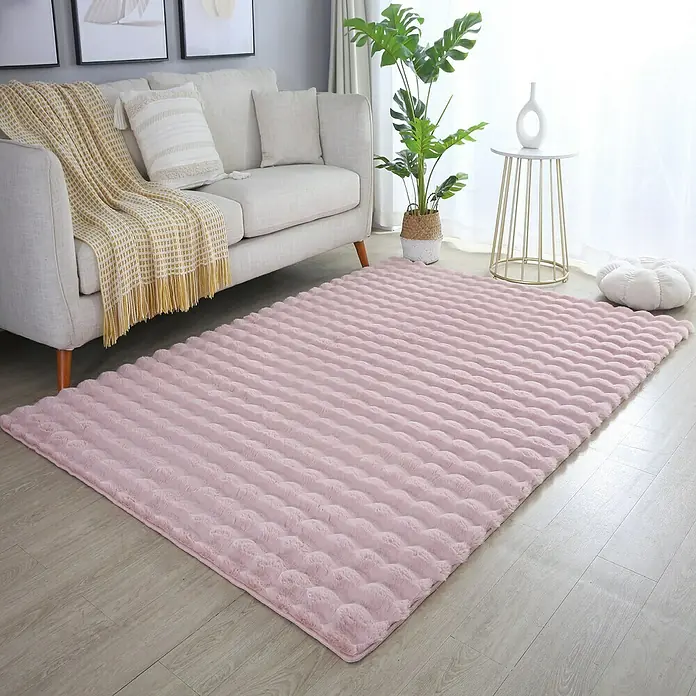 Fellteppich Ambiance - Rosa, 170 x 120 cm, 100 % Polyester - Ein weicher, rosa Teppich mit wellenförmiger Textur liegt in einem Wohnzimmer mit Sofa, Pflanze und Beistelltisch.