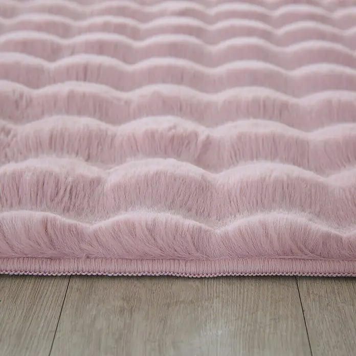 Fellteppich Ambiance - Rosa, 170 x 120 cm, 100 % Polyester - Nahaufnahme eines rosa, gewellten Teppichs auf einem Holzboden, der eine weiche und luxuriöse Textur zeigt.