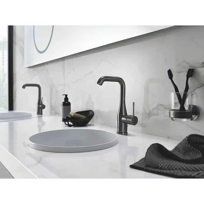 Grohe Essence New Waschtischarmatur L-Size - Mit Zugstange, Hard Graphite, Matt - Elegantes Badezimmer-Interieur mit schlankem, dunklem Wasserhahn, runden Waschbecken und stilvollem Zubehör auf einer Marmorplatte.