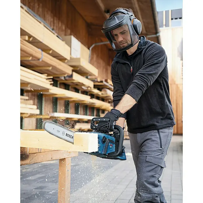 Bosch Professional AMPShare 18V Akku-Kettensäge GKE 18V-40 - 18 V, Li-Ionen, Ohne Akku, Schwertlänge: 40 cm - Ein Mann mit Gesichtsschutz sägt ein Holzstück mit einer blauen Bosch Akku-Kettensäge. Sägespäne fliegen im Vordergrund.