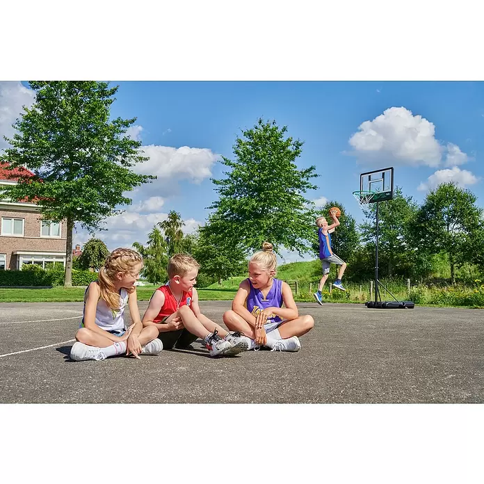 Salta Basketballkorb Dribble - Schwarz - Tragbarer Basketballkorb im Freien. Ein Junge springt zum Wurf, drei Kinder sitzen zuschauend auf dem Platz.