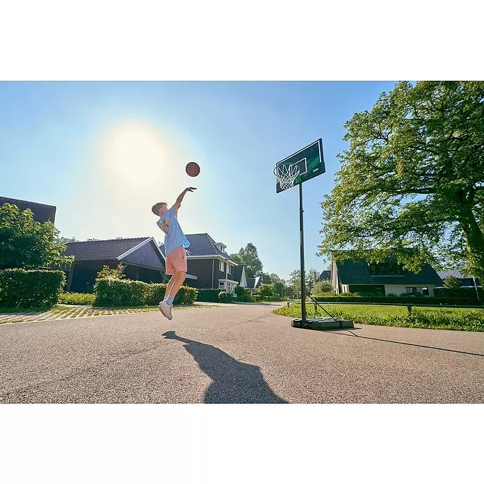 Salta Basketballkorb Forward - L x B x H: 172 x 110 x 362 cm, Schwarz - Ein Junge springt, um einen Basketball in einen tragbaren Korb mit schwarzer und grüner Rückwand auf einem Außenplatz zu werfen.