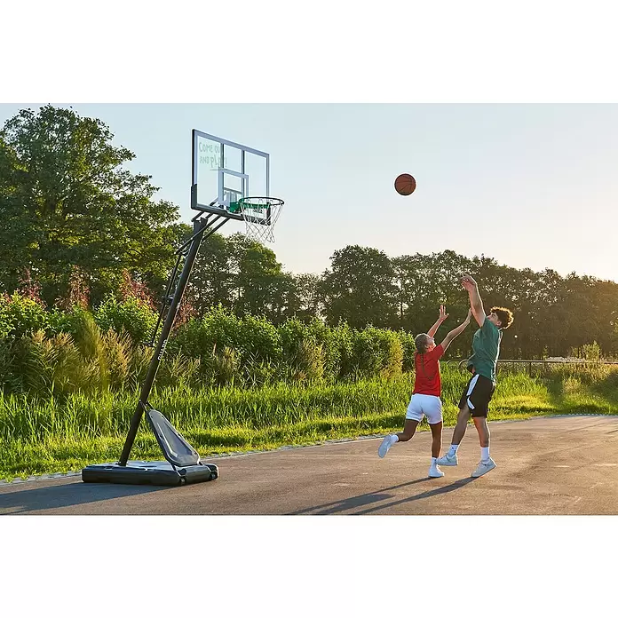 Salta Basketballkorb Guard - Schwarz, Höhe: 371 cm - Zwei Personen spielen Basketball mit einem tragbaren Korb. Er hat eine transparente Rückwand, grüne Akzente und einen dunklen Standfuß auf dem Freiplatz.