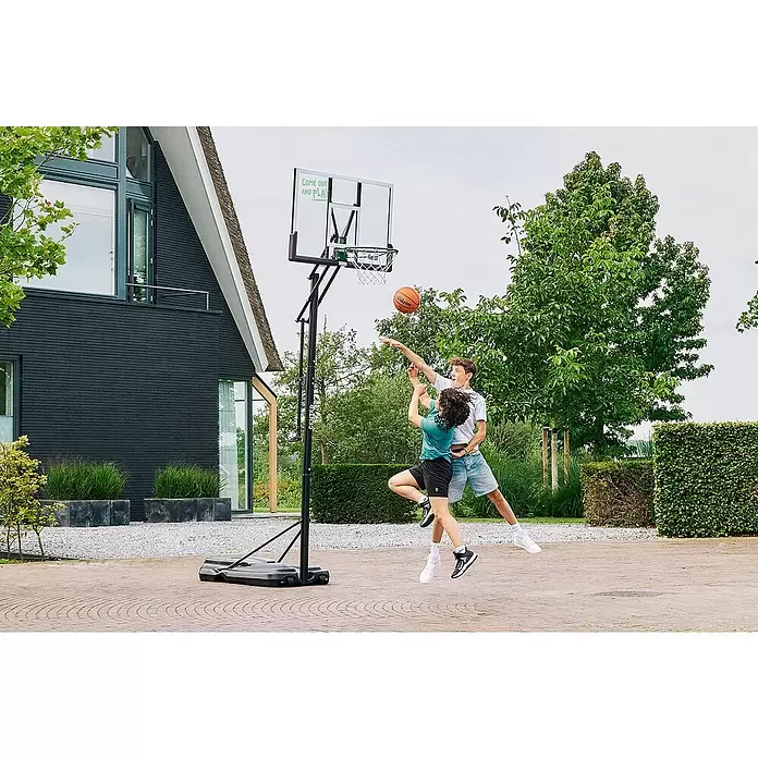 Salta Basketballkorb Center - 234 x 210 x 125 cm, Schwarz - Zwei Personen spielen Basketball mit einem schwarzen tragbaren Korb und transparenter Platte auf gepflastertem Außenbereich.