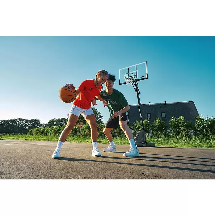 Salta Basketballkorb Guard - Schwarz, Höhe: 371 cm - Zwei junge Erwachsene spielen Basketball im Freien. Eine Person in Rot hält einen orangefarbenen Wilson-Basketball, während eine andere in Grün daneben lächelt.
