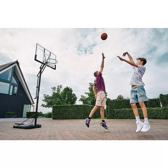 Salta Basketballkorb Center - 234 x 210 x 125 cm, Schwarz - Zwei Jungen spielen Basketball mit einem tragbaren Korb mit klarer Rückwand und grünem Ring, wobei einer einen Ball wirft und der andere blockiert.