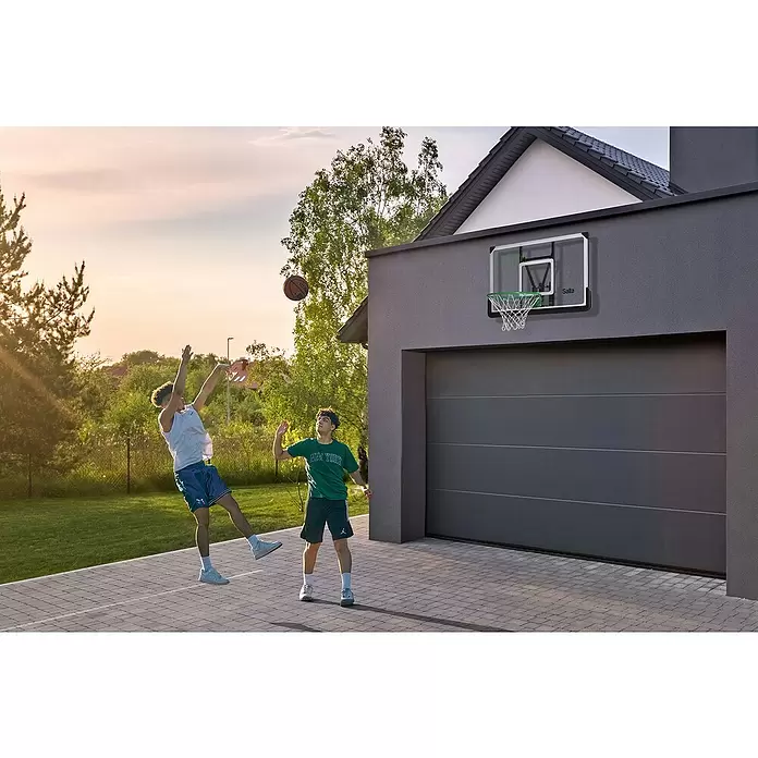 Salta Basketballkorb - 115 x 75 x 80 cm, Schwarz - Ein Mini-Basketballkorb mit klarer Rückwand und grünem Ring an einer dunkelgrauen Garagenwand, mit zwei Jungen beim Basketballspielen.