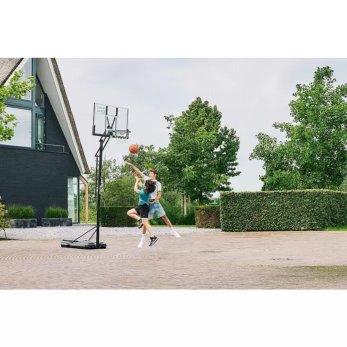 Salta Basketballkorb Center - 234 x 210 x 125 cm, Schwarz - Zwei Jungen spielen Basketball mit einem schwarzen, transparenten, tragbaren Basketballkorb auf einer Einfahrt. Der Korb hat eine Rückwand mit Text.