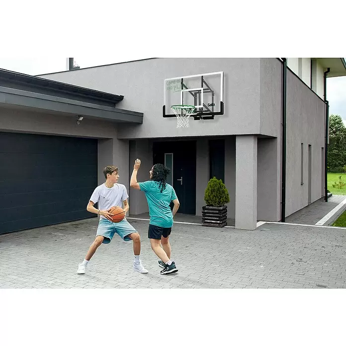 Salta Basketballkorb Guard - 138 x 86 x 128 cm, Schwarz - Basketballkorb mit transparenter Platte, schwarzem Rahmen, grünem Ring und weißem Netz an einer Hauswand, zwei Personen spielen Basketball.