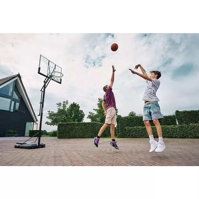 Salta Basketballkorb Center - 234 x 210 x 125 cm, Schwarz - Schwarzer, mobiler Basketballkorb mit transparenter Platte und grünen Akzenten, von zwei Jungen beim Spielen im Freien benutzt.