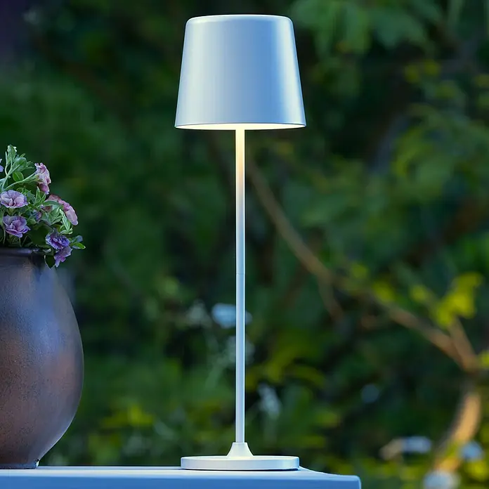 Brilliant Akku-Tischleuchte Kaami - 2 W, Weiß, Warmweiß - Elegante weiße Tischleuchte mit warmem Lichtschein, ideal für stimmungsvolle Abende im Freien. Dekorativ neben einer Blumenvase.