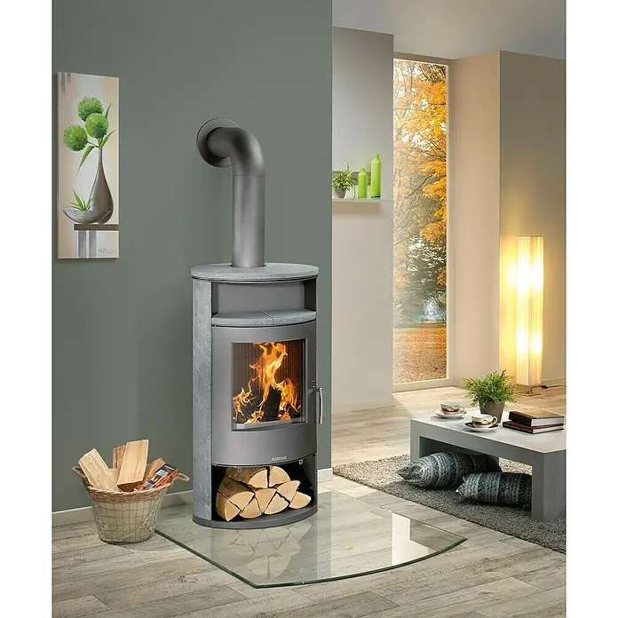 Justus Kaminofen Island Aqua - 10 kW, Raumheizvermögen: 200 m³, Material Verkleidung: Speckstein, Gussgrau - Ein Justus Kaminofen mit Glasbodenplatte und Holzkorb in einem modern eingerichteten Wohnzimmer mit herbstlichem Ausblick.