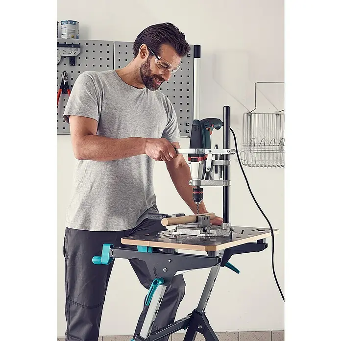 Wolfcraft Bohrmaschinenhalter - L x B x H: 160 x 230 x 500 mm, Hub: 60 mm - Ein Mann mit Schutzbrille benutzt eine Wolfcraft Bohrmaschine mit Ständer zum Bohren von Holz auf einer Werkbank.