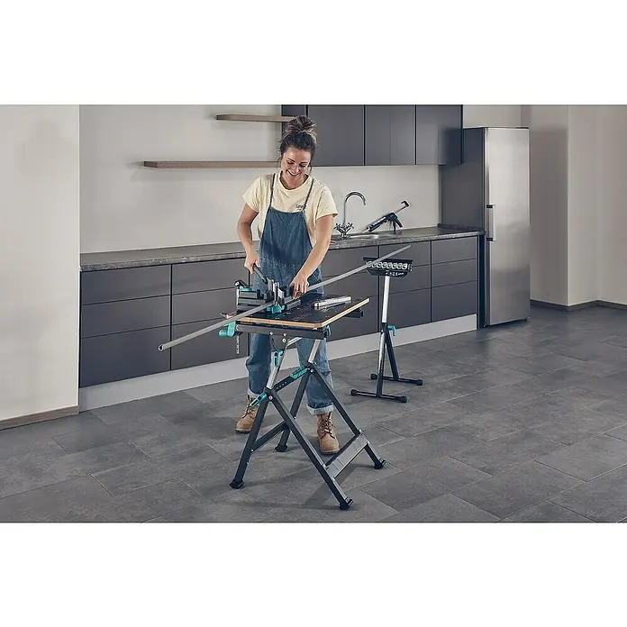 Wolfcraft Multifunktions-Auflagebock - Arbeitshöhe: 630 mm - 1.000 mm, Belastbarkeit: 50 kg - Eine Frau benutzt einen Wolfcraft Werkzeugtisch mit einer Metallsäge, um ein Werkstück zu bearbeiten. Im Hintergrund eine Küchenzeile.