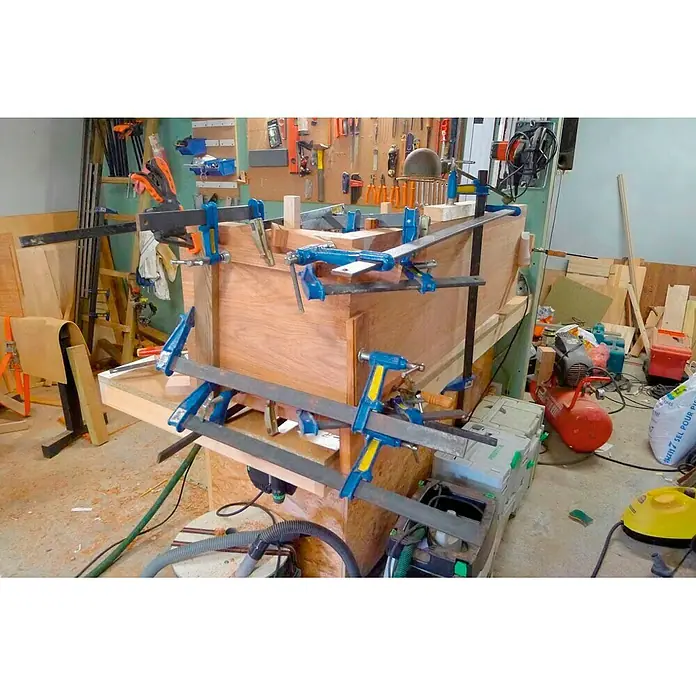 Piher Sargento F-40 - Anchura de sujeción: 400 mm, Fuerza de tensión: 9.000 N, Apertura máxima: 120 mm - Una pieza de madera asegurada con una serie de abrazaderas azules y amarillas en un taller. Precisión y estabilidad son clave.