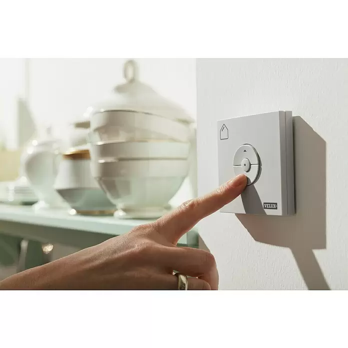 Velux Funk-Wandschalter Sonnenschutz KLI 312 - Passend für: Innen liegende VELUX Solar- & Elektro-Sonnenschutzprodukte (io-homecontrol®) - Eine Hand drückt ein hellgraues VELUX Bedienfeld mit Haus-Symbol und Richtungstaste.