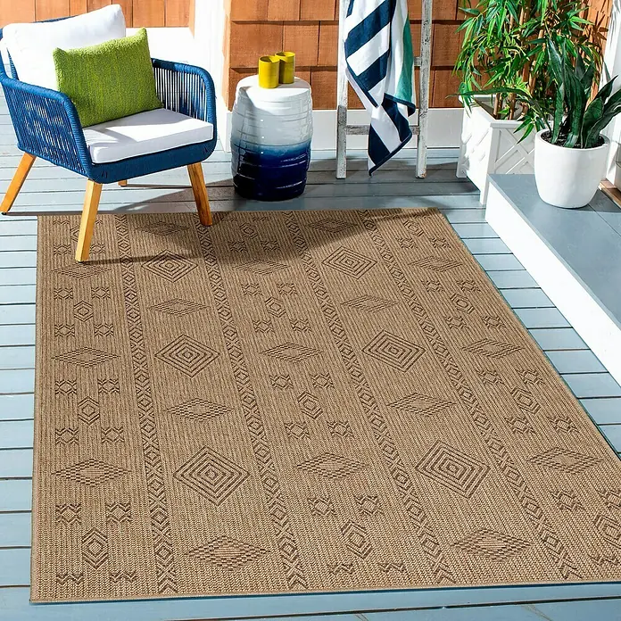 Outdoor-Teppich Dhaka III - Schwarz, 290 x 200 cm, 100 % Polypropylen - Ein brauner Outdoor-Teppich mit geometrischen Mustern liegt auf einer Terrasse, neben einem blauen Stuhl und Pflanzen.