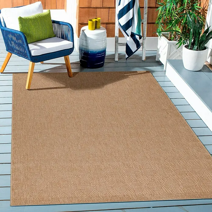 Outdoor-Teppich Dhaka VI - Beige, 150 x 80 cm, 100 % Polypropylen - Ein Outdoor-Teppich in natürlicher Farbe liegt auf einer blauen Holzterrasse neben einem Stuhl, der mit weißen Kissen und einem grünen Kissen ausgestattet ist.