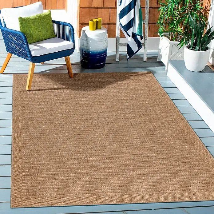 Outdoor-Teppich Dhaka II - Beige, 230 x 160 cm, 100 % Polypropylen - Ein Outdoor-Teppich in Braun liegt auf einer Holzterrasse neben einem blauen Sessel, der mit einem grünen Kissen dekoriert ist.