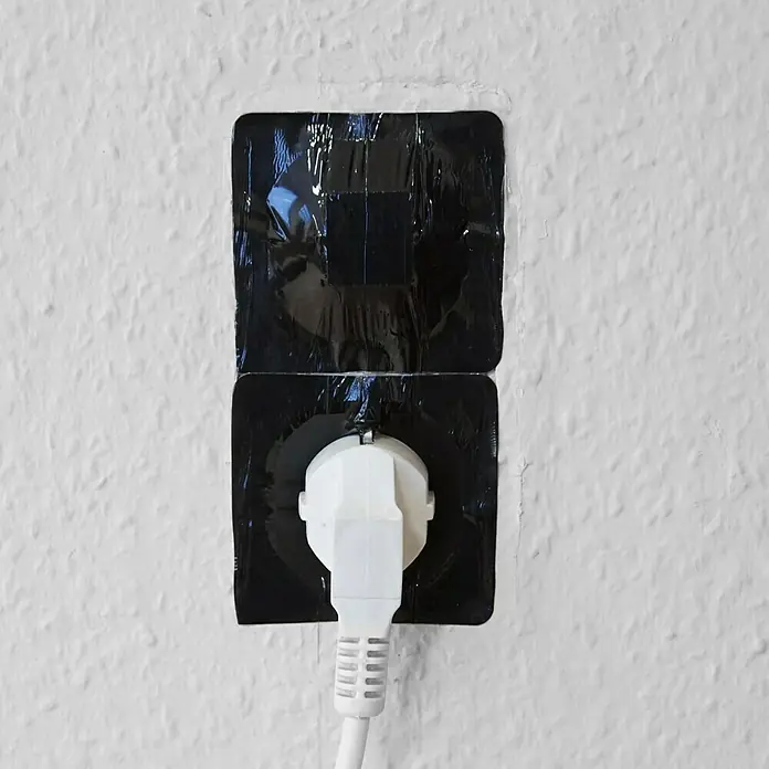 Mako Abdeckband - 70 x 70 mm - Das Bild zeigt eine Steckdose mit schwarzem Isolierband abgedeckt, in die ein weißer Stecker eingesteckt ist. Die Wand ist weiß und strukturiert.