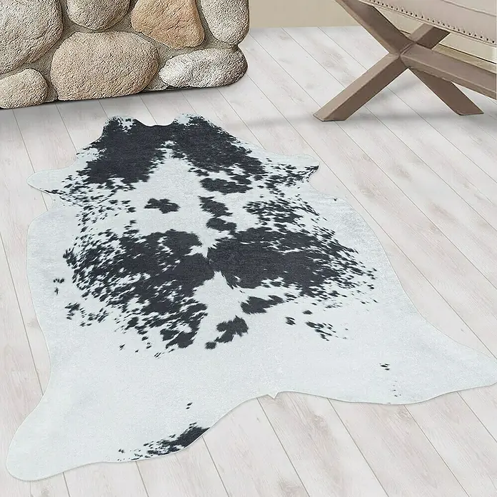 Flachgewebeteppich Etosha - Kuh, Schwarz/Weiß, 200 x 150 cm, 100 % Polyester - Ein Kuhfellteppich mit schwarzen Flecken liegt auf einem hellen Holzboden, neben einem Steinkamin und einem Holzbeistelltisch.