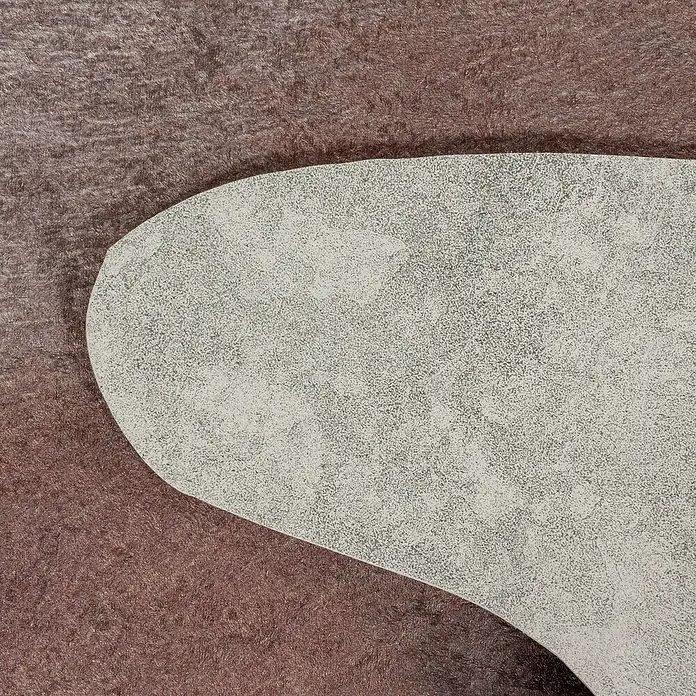 Flachgewebeteppich Etosha - Kuh, Braun, 200 x 150 cm, 100 % Polyester - Nahaufnahme einer strukturierten, hellgrauen Oberfläche, die einen Teil eines Produkts oder Designs hervorhebt, vor einem weichen, braunen Hintergrund.