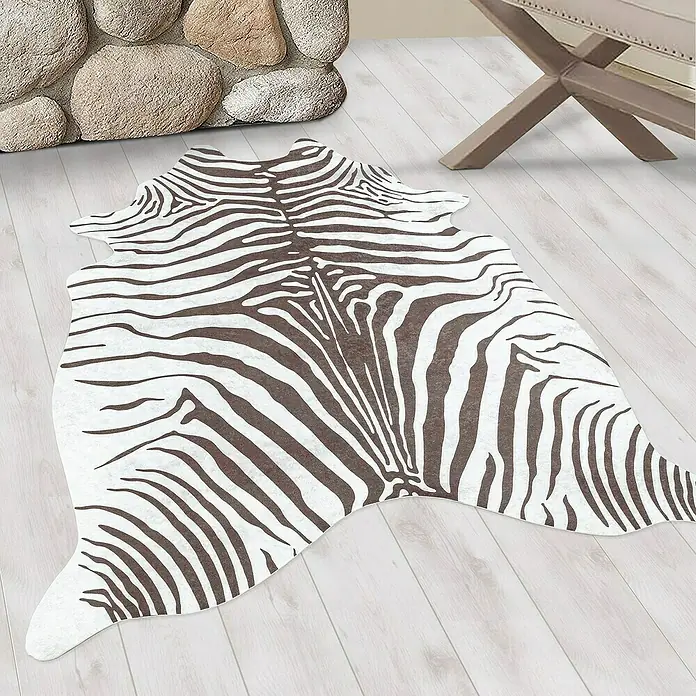 Flachgewebeteppich EtoshaZebra, Braun, 200 x 150 cm, 100 % Polyester - Ein Kuhfellteppich mit Zebra-Muster liegt auf einem hellen Holzboden neben einem Steinkamin und einem Hocker.