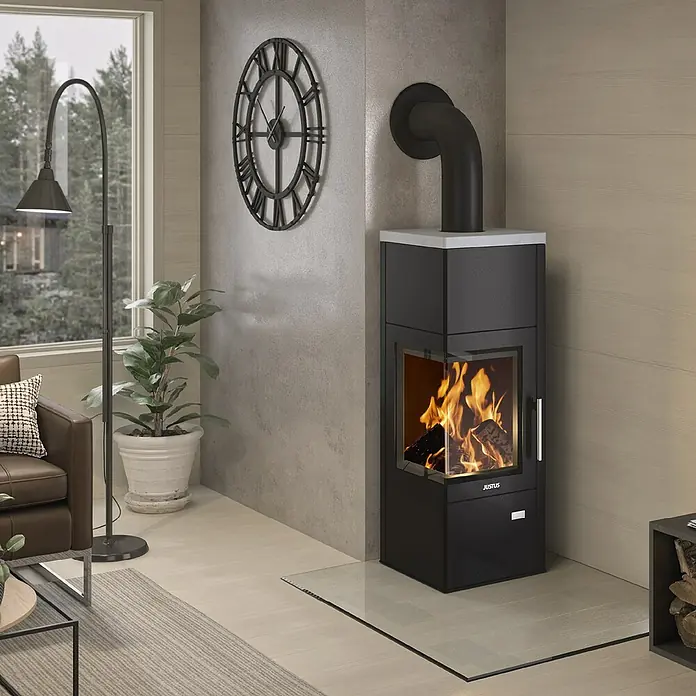 Justus Kaminofen Diego wärmePLUS - 6 kW, Raumheizvermögen: 115 m³, Material Verkleidung: Speckstein, Schwarz/Speckstein - Ein moderner Justus Kaminofen mit Feuer, platziert in einem stilvollen Wohnzimmer mit Uhr, Lampe und Pflanze.