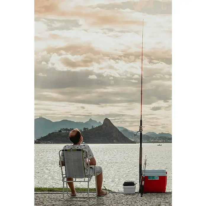 Bote hinchable para pesca - Apto para: 1 persona, Verde - Un hombre está sentado en una silla junto al agua, con una caña de pescar y una nevera portátil a su lado, disfrutando de la vista de las montañas.