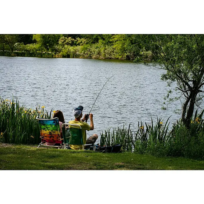 Bote hinchable para pesca - Apto para: 1 persona, Verde - Relajación junto al lago: Un hombre pesca con su caña mientras una mujer se relaja en una silla colorida junto al agua.