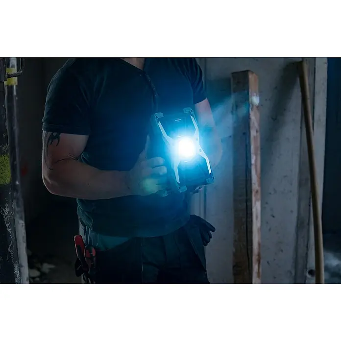Brennenstuhl Mobiler LED-Strahler Rufus - Akkubetrieben, 30 W, L x B x H: 5 x 14 x 21 cm, IP65 - Ein Mann hält eine helle LED-Arbeitsleuchte in einem dunklen Raum, die das Arbeitsumfeld erhellt. Das Gerät ist robust und ideal für Baustellen.