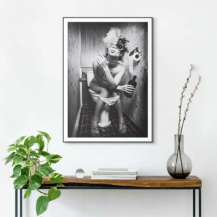 Reinders Bild Slim Frame Black - Toilet Picture, B x H: 50 x 70 cm - Schwarzes gerahmtes Poster an einer weißen Wand, das eine Frau auf einer Toilette beim Rauchen und Trinken zeigt, auf einem Holztisch platziert.