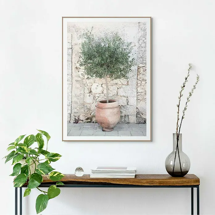 Reinders Bild Slim Frame Wood - Olive Tree, B x H: 50 x 70 cm - Gerahmtes Wandbild mit Olivenbaum im Topf vor einer Steinwand, platziert auf einem Holztisch mit Dekorationen.