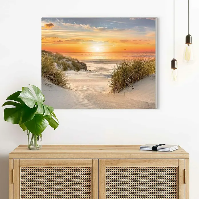 Reinders Decopanel - Beautiful Dunes, B x H: 50 x 40 cm - Ein gerahmtes Strandbild mit Sonnenuntergang hängt über einer Kommode mit Rattandetails, daneben eine Zimmerpflanze und Notizbücher.