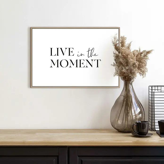 Reinders Bild Slim Frame Wood - Live in the Moment, B x H: 30 x 20 cm - Ein gerahmtes Kunstwerk mit dem Schriftzug "Live in the Moment" hängt über einem eleganten Tisch mit stilvoller Dekoration.