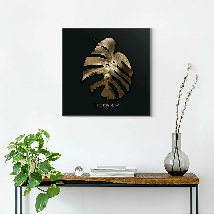 Reinders Bild Slim Frame Black - Monstera Gold, B x H: 50 x 50 cm - Ein gerahmtes Kunstwerk mit einem goldenen Philodendronblatt auf schwarzem Hintergrund, das an einer weißen Wand hängt.