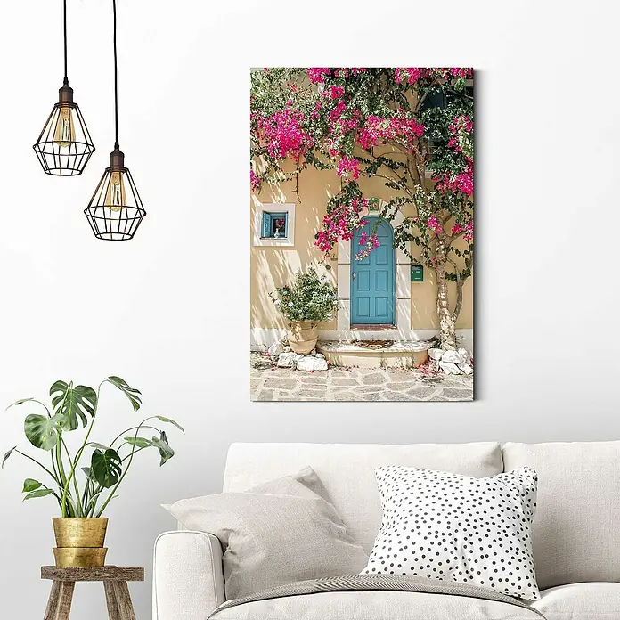 Reinders Decopanel - Mediterranean Door, B x H: 60 x 90 cm - Das Bild zeigt ein dekoratives Wandbild mit einem blauen Hauseingang, umrankt von rosa Blüten, über einem weißen Sofa im Wohnzimmer.