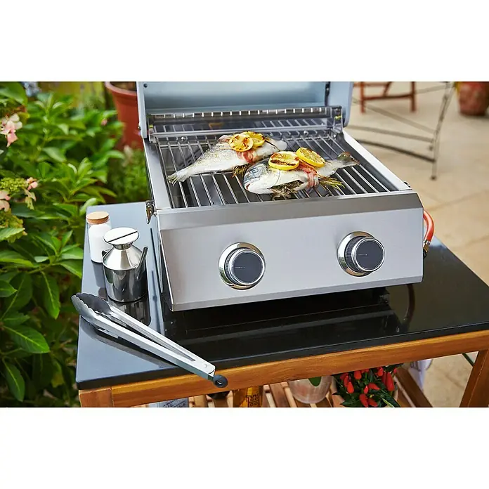 Grillstar Barbacoa de gas de mesa Atlanta 200 T - Superficie de la parrilla (An x Pr): 40 x 33 cm, 4,4 kW, Número de quemadores: 2 - Parrilla con pescado: Parrilla de gas compacta con dos pescados adornados con limones en las rejillas, en medio de una exuberante vegetación.