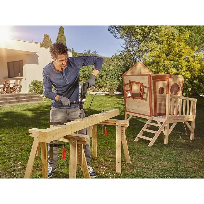 Bosch Advanced Schlagbohrmaschine AdvancedImpact 900 - 900 W, Schnellspannbohrfutter - Mann bohrt mit einer Bohrmaschine ein Holzbrett im Freien. Im Hintergrund steht ein Spielhaus.