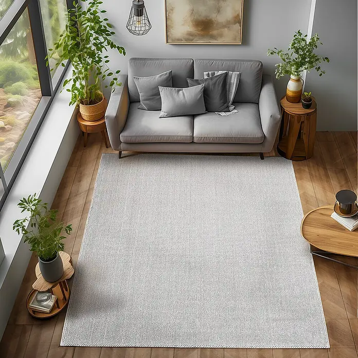 Flachgewebeteppich Loom - Silber, 230 x 160 cm, 100 % Polypropylen - Ein hellgrauer Teppich liegt in einem Wohnzimmer unter einem grauen Sofa, umgeben von Holzböden und Pflanzen.