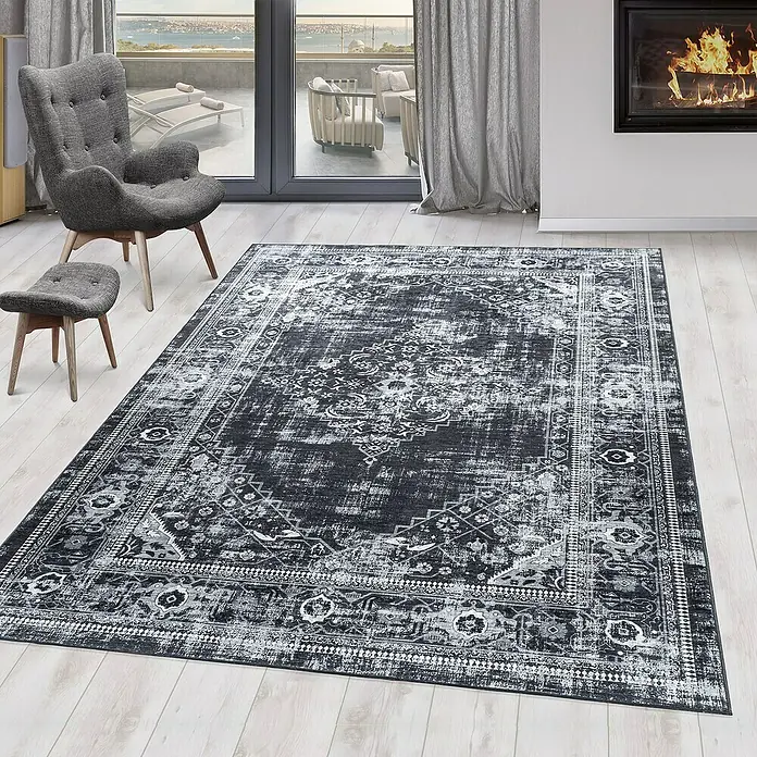 Flachgewebeteppich Fiesta - Schwarz/Grau, 290 x 200 cm, 100 % Polyester - Teppich mit kompliziertem Muster in einem Wohnzimmer mit Sessel und Kamin, der dem Raum einen Hauch von Eleganz verleiht.