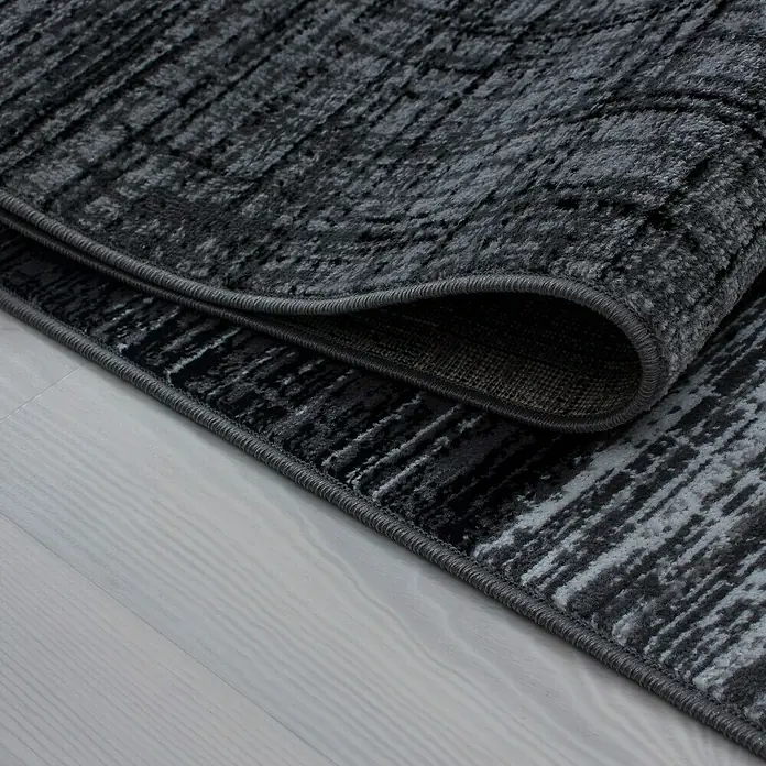 Kurzflorteppich Plus - Anthrazit/Grau, 370 x 280 cm, 100 % Polypropylen - Nahaufnahme eines grauen Teppichs mit abstraktem Muster und umgeschlagener Ecke, der die Textur und das Design hervorhebt.