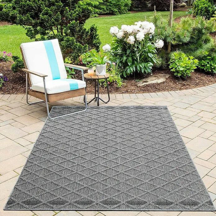 Outdoor-Teppich Patara V - Grau, 250 x 80 cm, 100 % Polypropylen - Ein grauer Teppich mit einem Rautenmuster liegt auf einer Terrasse neben einem Stuhl und einem Beistelltisch.