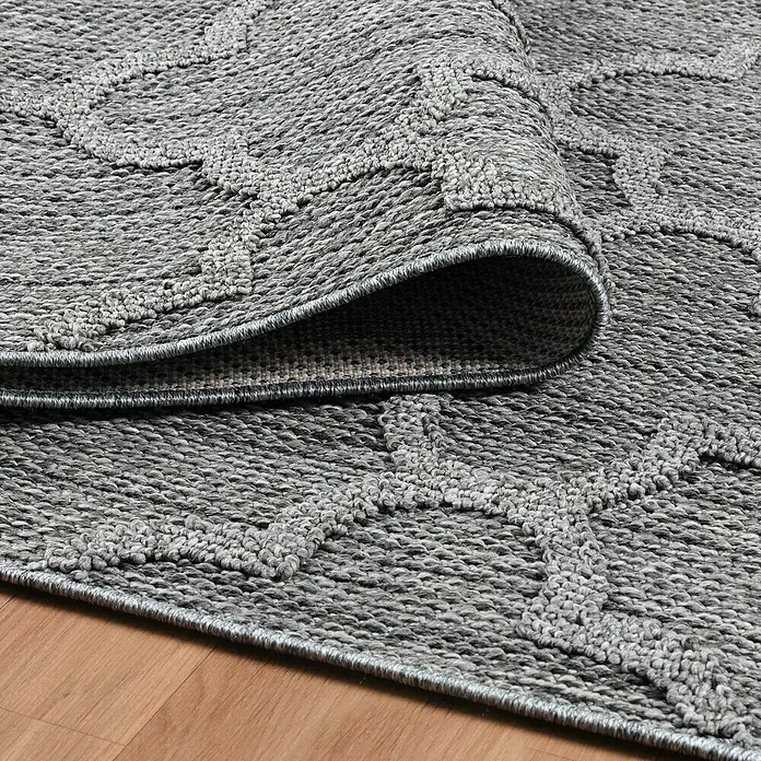 Outdoor-Teppich Patara IV - Grau, 230 x 160 cm, 100 % Polypropylen - Nahaufnahme eines grauen Teppichs mit einem gewebten Muster, der teilweise aufgerollt ist und die Textur zeigt.