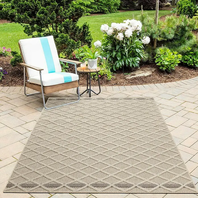 Outdoor-Teppich Patara V - Beige, 200 x 140 cm, 100 % Polypropylen - Ein gemusterter Teppich im Freien liegt auf einer Terrasse neben einem Stuhl und einem Beistelltisch, umgeben von üppigem Grün.